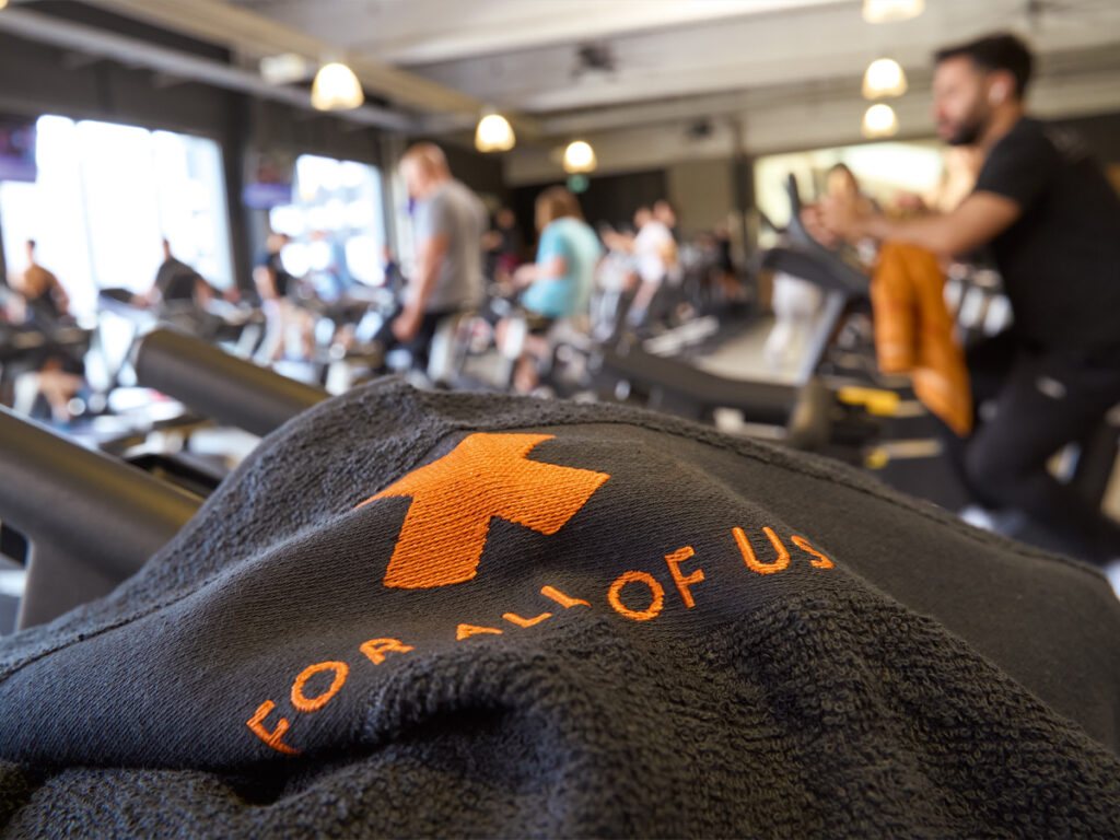 Im Vordergrund ein schwarzes Handtuch, im Hintergund trainierende Menschen im Fitnessstudio